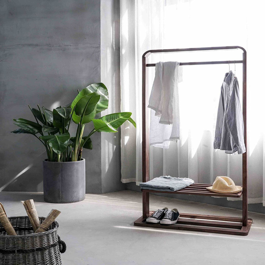 Room with a wooden portable hanger for clothes and shoes. On the left hand has a big grey plant pot, next to it a a window with white shimmer curtains. In front of the window is the portable hanger.